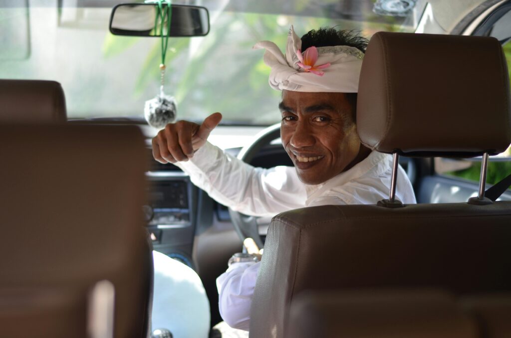 A Balinese man wearing traditional attire gives a thumbs up and smiles warmly from inside a car in Ubud, Bali.